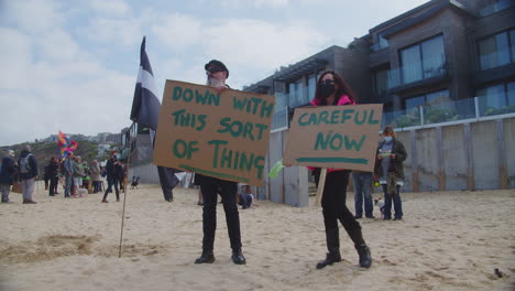 Demonstrating-against-the-Carbis-Bay-Hotel,-two-protesters-holding-signs,-stood-on-the-beach,-Cornwall