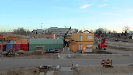 Aktivitäten-Auf-Der-Baustelle-Mit-Einem-Hebefahrzeug-Und-Einem-Anhängerwagen,-Die-Einen-Teil-Eines-Provisorischen-Bürogebäudes-Anheben-Und-An-Einen-Anderen-Ort-Verlegen,-Und-Im-Vordergrund-Das-Abladen-Schwerer-Maschinen