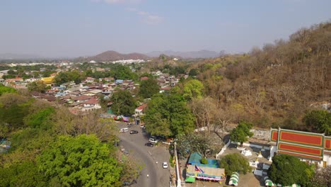 Wat-Phra-Phutthabat,-Saraburi,-Thailand,-ascending-aerial-footage-revealing-the-road-with-cars,-Thai-Flag,-entrance-into-the-teple,-community-and-houses,-hills-and-the-horizon,-blistering-summer-day