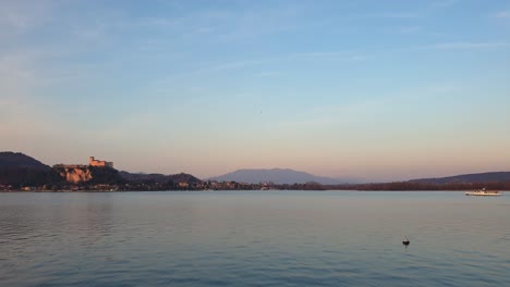Die-Fähre-Fährt-Bei-Sonnenuntergang-Auf-Dem-Lago-Maggiore-Mit-Der-Festung-Angera-Im-Hintergrund