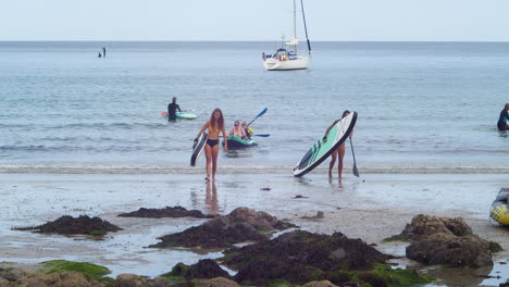Female-Tourists-In-Bikinis-Walking-At-The-Beach-With-Paddleboards-And-Paddles-In-Coverack,-Lizard-Peninsula,-England