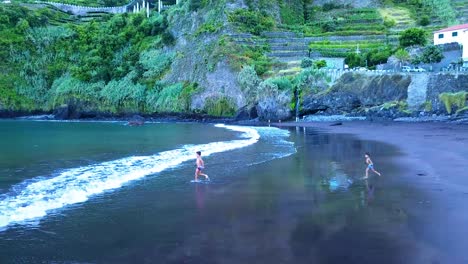 Two-children-play-and-run-into-the-sea-on-volcanic-black-sand-of-Seixal-beach-in-Madeira,-Portugal