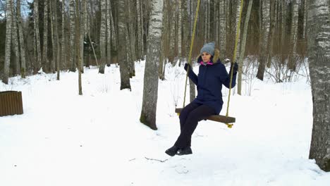 A-young-caucasian-girl-swinging-on-winter-forest-in-snow-covered-winter-birch-forest-at-sunny-day,-alone-in-frosty-weather,-rotating-to-the-right-gimbal-shot