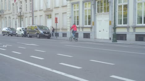 Cyclist-in-red-jacket-on-foldable-city-bike-cycling-in-bike-lane-in-rich-city-street-in-Brussels,-Belgium-with-Belgian-flags-in-the-distance