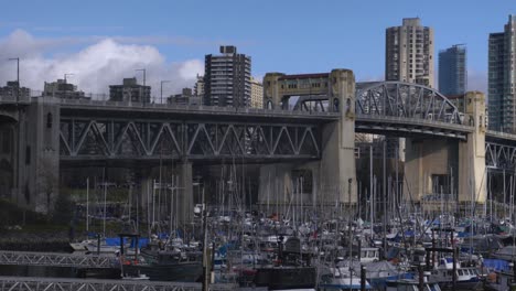 Panning-across-bridge-over-boat-dock-and-marina-small-boats-anchored-at-waterfront-cityscape-high-rise-buildings-and-a-bridge-in-the-background