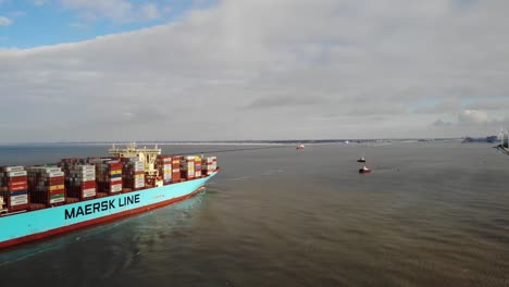 Buque-Contenedor-Gigante-Cargado-Con-Contenedores-En-Cubierta-Que-Ingresa-Al-Puerto-De-Rotterdam-Con-Dos-Pequeños-Remolcadores-Esperando-Para-Ayudar-Al-Buque-A-Atracar-En-Un-Día-Tranquilo-Parcialmente-Nublado