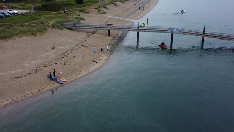 Paddle-boarding-couple-in-preparation-on-sandy-British-beach-as-jet-ski-appears-under-jetty-aerial-view