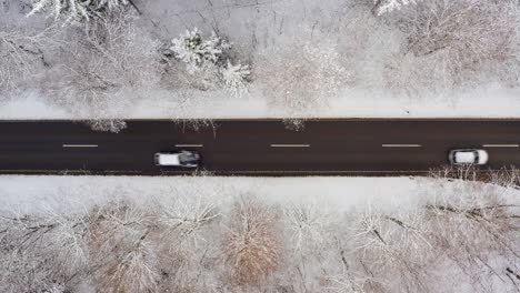 Two-cars-are-driving-over-a-winter-road-from-left-to-right,-top-down-drone-shot