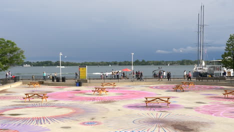 People-Strolling-By-Waterfront-Park-In-Toronto,-Canada-With-Colorful-Arts-On-The-Ground-In-Foreground
