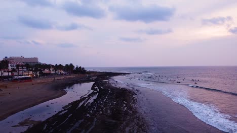 Unrecognizable-big-group-of-people-with-surfboards-in-Tenerife-beach-surfing-at-sunset,-aerial-drone-shot
