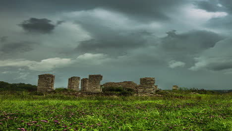 Timelapse-shot-of-dark-cloud-movement-over-abandoned-stone-structure-surrounded-by-green-grasslands-during-sunset
