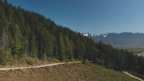 Red-jeep-SUV-off-road-vehicle-driving-on-dirt-logging-road-approaching-trees-on-mountain-forest-beautiful-scenery-blue-sky-snow-covered-peaks-wildflowers-in-spring-Aerial-follow-very-wide-panorama