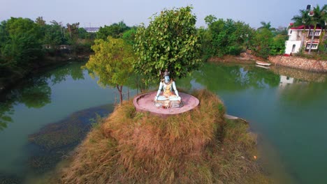 Estatua-Azul-Del-Dios-Hindú-Shiva-Sentado-Con-Las-Piernas-Cruzadas-En-Un-Islote,-Rodeado-Por-Un-Lago-Verde-Azulado-Y-Una-Mansión-Blanca-Al-Fondo,-En-Vadodara,-India