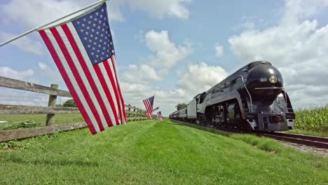A-Low-Angel-of-a-Steam-Engine-and-Passenger-Coaches-Approaching-With-a-Row-of-American-Flags-lining-the-Approach-on-a-Beautiful-Day