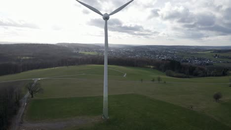 Fast-spinning-wind-turbine-surrounded-by-green-meadows-and-brown-patches-of-forest-underneath-a-cloudy-sky