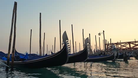 Low-angle-water-surface-view-of-row-of-docked-gondolas-at-sunset,-Venice-in-Italy