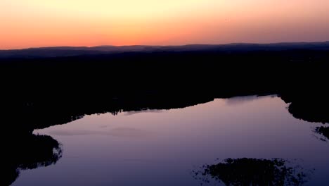 Luftaufnahme-über-Einem-Ruhigen,-Glatten-See-Mit-Violettem-Reflektiertem-Licht-Vor-Orangefarbenem-Sonnenuntergangshimmel,-Der-Von-Wäldern-Umrahmt-Wird