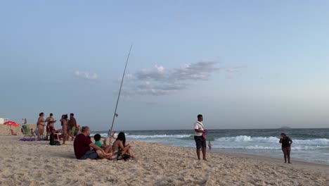 Family-around-fishing-rod-at-Ipanema-beach-with-soft-sunset-golden-hour-sunlight-and-fluffy-clouds-above-the-waves-coming-in-on-the-well-treaded-sand