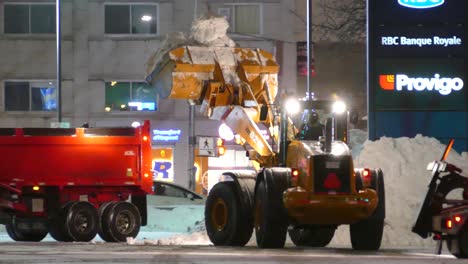 Máquina-Excavadora-De-Pala-Cargando-Camión-Con-Nieve-De-Trabajos-De-Limpieza-Por-La-Noche,-Paisaje-Urbano