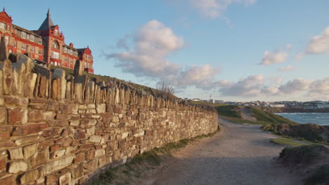 View-from-Coastal-Path-of-Beachfront-Headland-Hotel-and-Spa-Sunlit-at-Dusk-In-Newquay,-Cornwall,-United-Kingdom---low-wide-panning