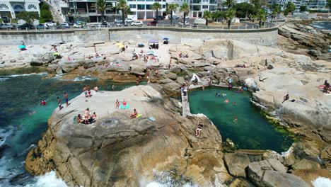 people-relaxing-and-sun-tanning-at-Saunders-Rocks-Beach-during-summer-in-Cape-Town,-aerial
