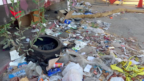 Heap-of-Trash-Lying-on-Sidewalk-by-Graffiti-Wall-in-Daytime,-Rubber-Tire-and-Fast-Food-Wrappers-Littered-on-Pavement