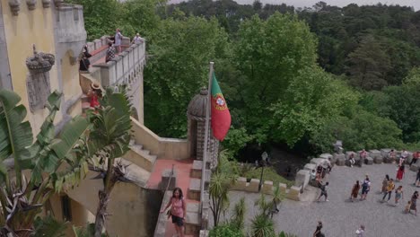 Tourists-roam-upper-levels-of-stunning-iconic-historic-Pena-Palace-with-Portugese-flag-waving-in-front-of-yellow-architecture-building,-Sintra,-Lisbon,-Portugal