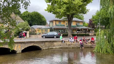 Familie-Sitzt-In-Der-Nähe-Einer-Brücke-Und-Beobachtet-Den-Windrausch-Des-Flusses,-Während-Radfahrer-Auf-Rennrädern-In-Bourton-On-The-Water,-Cotswolds,-England,-Großbritannien,-Vorbeifahren