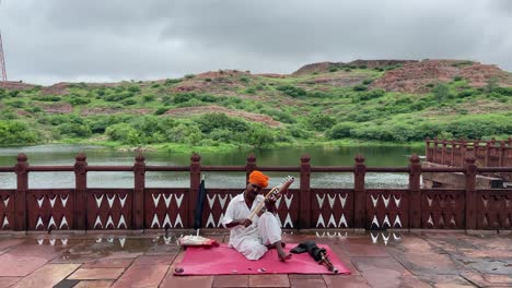 Static-shot-of-local-Indian-man-seated-playing-a-traditional-instrument-in-front-of-a-lake-in-an-idyllic-landscape