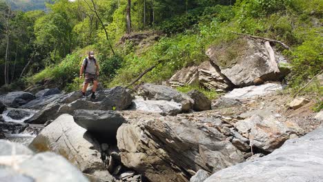 Man-walking-over-rocks-and-boulders-near-a-free-flowing-river-in-a-deep,-rugged-valley-wilderness-on-a-remote-tropical-island-landscape,-hiking-towards-camera
