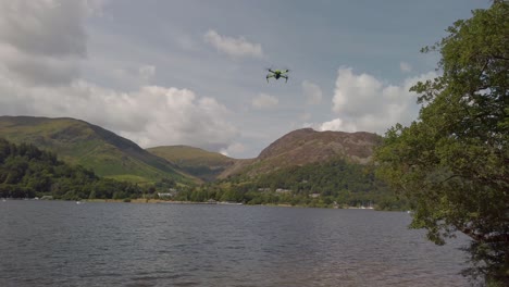 Small-drone-flying-over-Ullswater-in-the-Lake-District-National-Park-near-the-village-of-Glenridding,-with-the-Lakeland-Fells-and-a-cloudy-sky-in-the-background