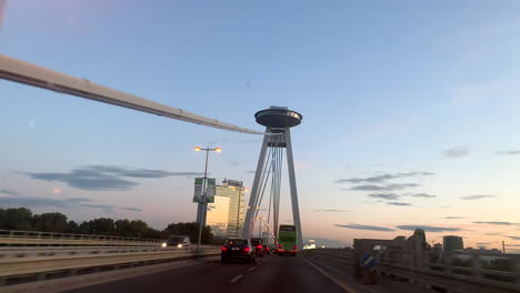 Drivers-perspective-driving-across-Danube-bridge-in-Bratislava,-Slovakia-beneath-famos-UFO-landmark-with-observation-deck-and-restaurant,-popular-tourist-destination