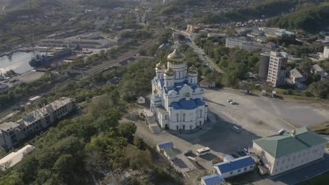 Slow-pivot-aerial-shot-of-an-orthodox-church-with-blue-roof-and-golden-domes,-located-on-top-of-the-hill-with-port-and-city-building-in-the-background,-on-a-bright,-clear,-sunny-day