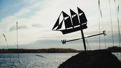 Metallic-Weather-Vane-On-A-Rock-By-The-Lake-With-Sunlight-And-Cloudy-Sky-In-Background-In-Rauma,-Finland---Medium-Shot