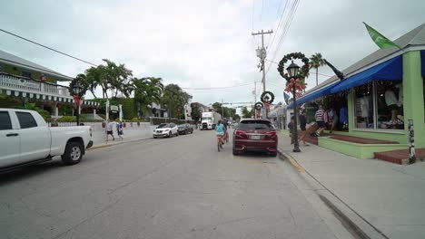 Key-West-Street-Mit-Straßenlaternen,-Die-Mit-Weihnachtskränzen-Geschmückt-Sind,-Und-Einem-Paar,-Das-Auf-Beachcruisern-Die-Straße-Entlang-Fährt