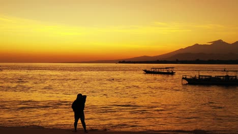 Silhouette-of-woman-greets-people-in-the-anchored-boats-on-calm-lagoon-reflecting-orange-sky-after-beautiful-sunset,-Bali