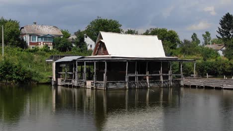 Low-Aerial-View-of-Rugged-Shack-on-a-River-With-a-Dock-and-Houses-in-Background-on-a-Sunny-Day