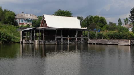 Low-Aerial-View-of-Rugged-Shack-on-a-River-With-a-Dock-and-Houses-in-Background-on-a-Sunny-Day