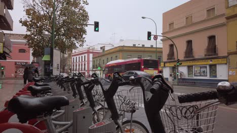 Bus-Und-Autos-Fahren-Im-Verregneten-Stadtpanorama-An-Einer-Reihe-Von-Leihfahrrädern-Vorbei