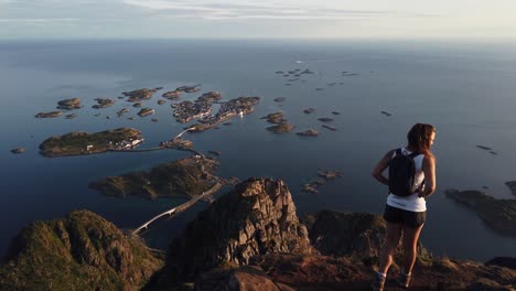 A-young-woman-is-standing-with-her-backpack-and-hiking-boots-on-top-of-Festvågtind-mountain,-overlooking-the-village-Henningsvaer-in-the-warm-midnight-sun-during-summer-in-Lofoten,-Norway