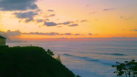 HD-Hawaii-Kauai-slow-motion-static-elevated-wide-shot-of-ocean-waves-crashing-in-from-right-to-left-with-a-person-on-a-lookout-point-in-distance-looking-at-a-beautiful-orange-sky-just-after-sunset