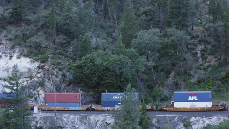 Cargo-train-carrying-shipping-containers-travels-slowly-through-the-mountains,-rising-aerial,-side-view,-flying-over
