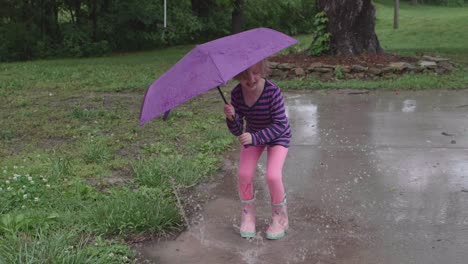 Young-Caucasian-girl-wearing-rain-boots-and-striped-shirt-holding-purple-umbrella-jumps-up-and-down-outside-splashing-water-in-puddle,-slow-motion-handheld-pan-up