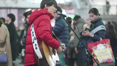 Un-Guitarrista-Japonés-Tocando-En-La-Calle-Fuera-De-La-Estación-De-Shinjuku-En-Tokio,-Japón---Toma-De-Cámara-Lenta-De-Pedestal-Hacia-Arriba