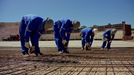 Men-work-in-a-line-as-they-ties-wire-around-a-grid-of-rebar-before-pouring-the-cement-foundation