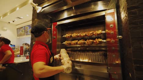 Rotisserie---Restaurantmitarbeiter-Mit-Gesichtsmaske-Und-Gesichtsschutz,-Der-Ganze-Hähnchen-Auf-Spieße-Im-Grill-Legt