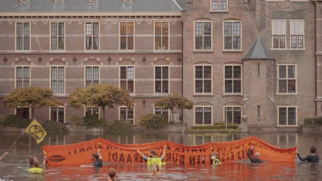 Demonstranten-Mit-Orangefarbenem-Banner-Gegen-Den-Klimawandel-Schwimmen-Im-Hofvijver-Teich,-Wasser-Vor-Dem-Niederländischen-Parlament,-Tweede-Kamer-Im-Binnenhof-Gebäude-–-Den-Haag,-Niederlande-–-Zeitlupe