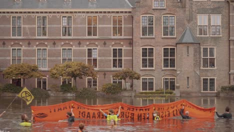 Manifestantes-Con-Pancarta-Naranja-Contra-El-Cambio-Climático-Nadando-En-El-Estanque-Hofvijver,-Agua-Frente-Al-Parlamento-Holandés,-Tweede-Kamer-En-El-Edificio-Binnenhof---La-Haya,-Países-Bajos---Cámara-Lenta