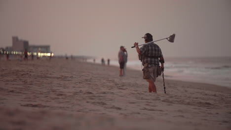 Man-with-metal-detector-searching-crowded-beach-at-sunset,-Slow-Motion