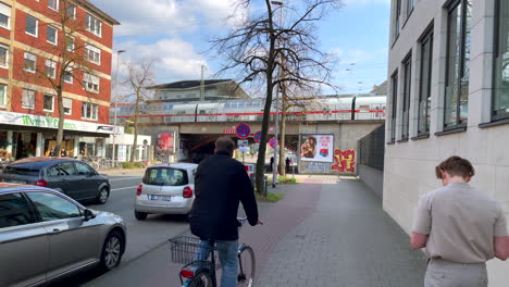 People-riding-bicycle,-pedestrian-walking-on-walkway-and-train-crossing-bridge-during-beautiful-sunny-day-in-german-large-city